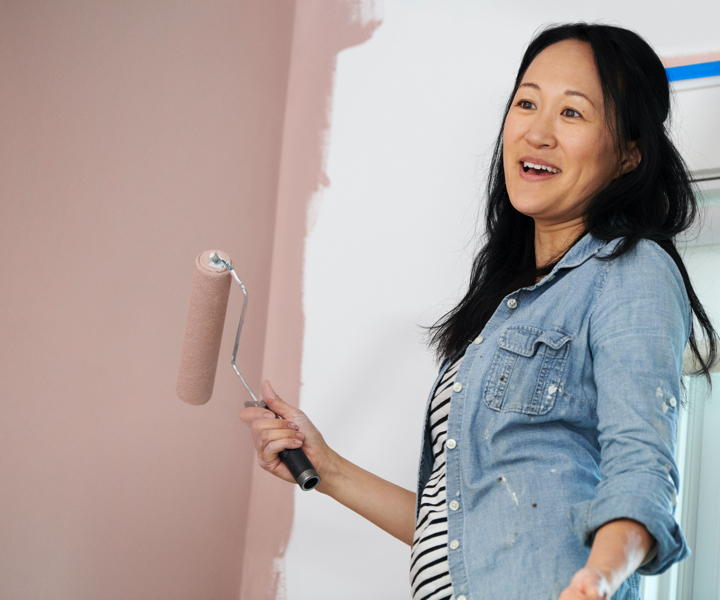 Smiling woman in denim and striped shirt holds roller as she is painting wall a rose-hued neutral.